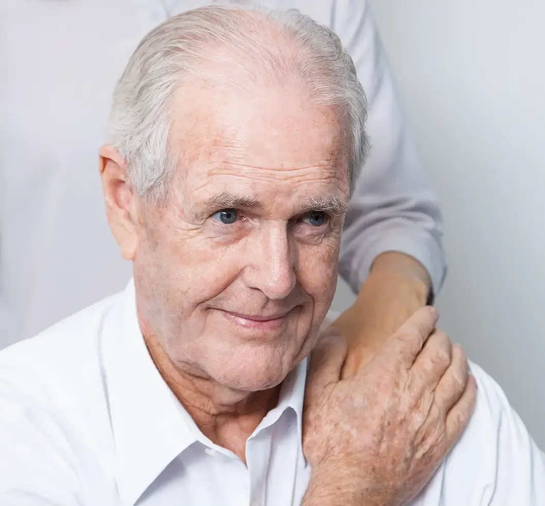 Ein älterer, lächelnder Patient in einem weißen Hemd wird fürsorglich betreut, eine schützende Hand liegt auf seiner Schulter.