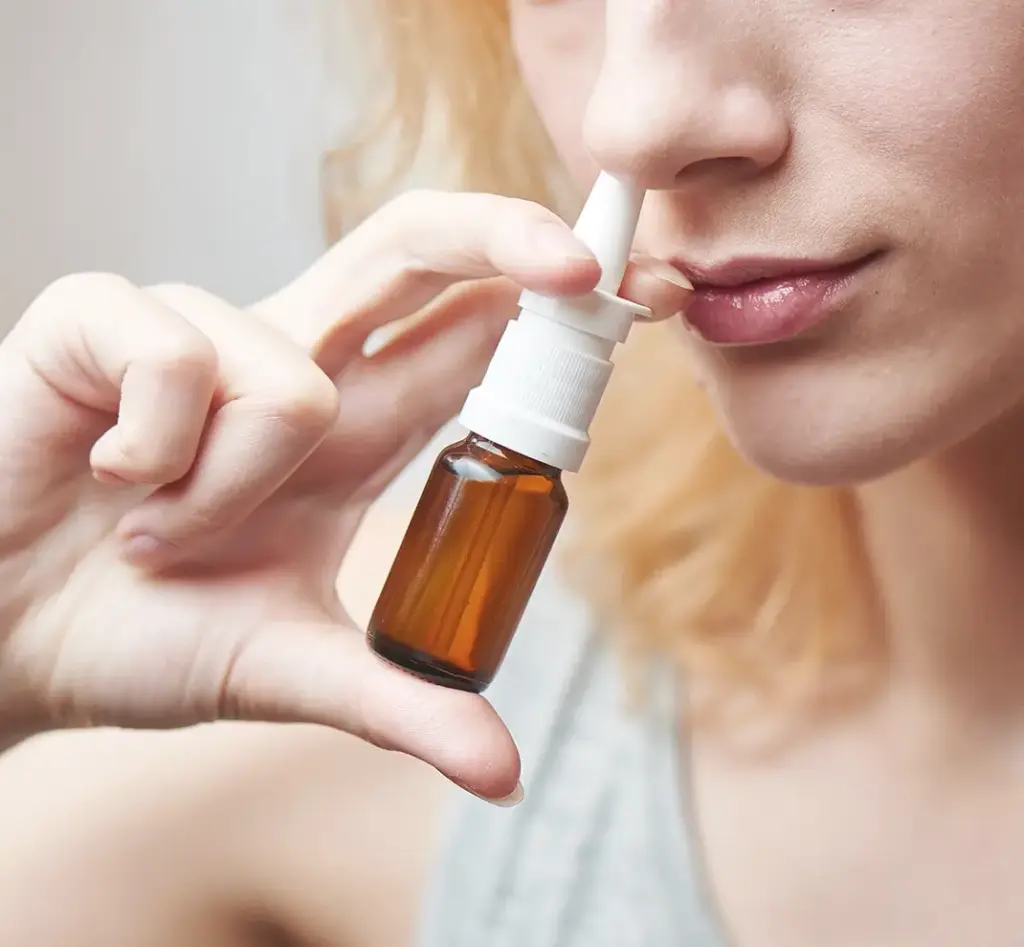 Eine junge Frau wendet ein Nasenspray in einer Glasflasche an – symbolisch für die medikamentöse Therapie von Schnupfen, Allergien und Nebenhöhlenentzündungen.