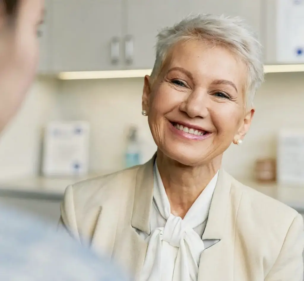 Eine elegant gekleidete, ältere Patientin blickt lächelnd und zufrieden im Gespräch zu einer Behandlerin in der HNO-Praxis Maren Festner.