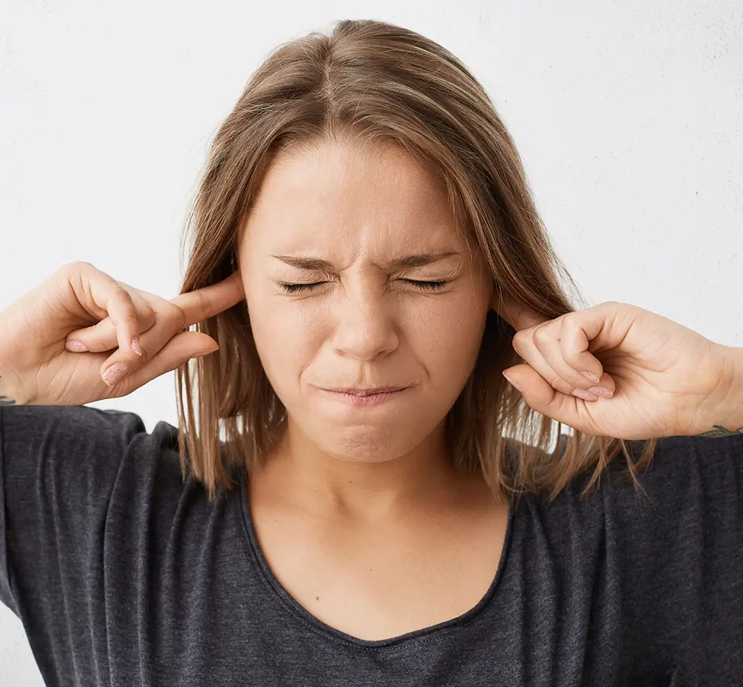 Eine junge Frau hält sich mit schmerzverzerrtem Gesicht die Ohren zu, symbolisch für Tinnitus.